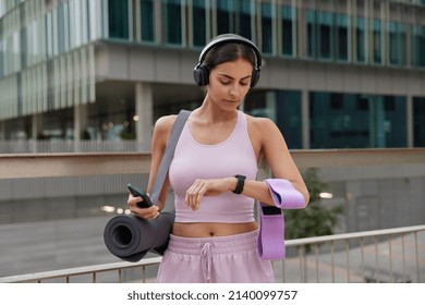 Outdoor shot of sporty fit woman poses with sport equipment checks time on smartwatch uses smartphone for tracking fitness results dressed in sportswear poses outdoors against urban background - Shutterstock ID 2140099757