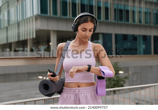 Outdoor shot of sporty fit woman poses with\
sport equipment checks time on smartwatch uses smartphone for\
tracking fitness results dressed in sportswear poses outdoors\
against urban\
background