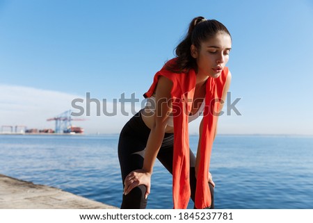 Outdoor shot of tired fitness woman panting and taking a breath after jogging, standing on a pier with sea behind her