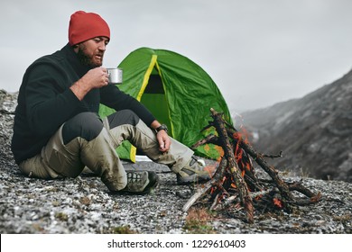 Outdoor shot of young breaded male drinking hot beverage in mountains. Traveler man in red hat, sitting near to camping tent, holding in hands a mug of tea after hiking. Travel, lifestyle. - Powered by Shutterstock