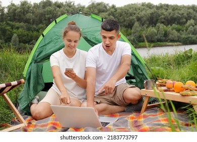 Outdoor Shot Of Young Family Sitting On Ground Near Tent And Using Laptop, Man Pointing At Notebook Screen With Shocked Expression, Woman Holding Cell Phone And Smiling.