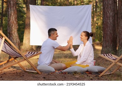 Outdoor Shot Of Young Positive Couple Sitting In The Forest With Overhead Projector With Empty White Screen With Advertisement Area, Family Holding Hands Or Giving High Five.