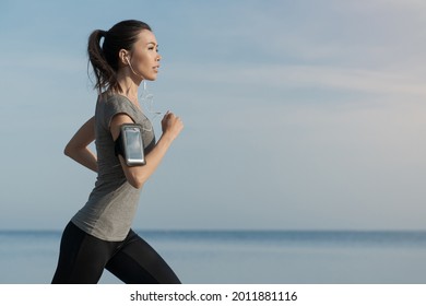 Outdoor shot of young sports asian woman running along the sea shore in morning. Female athlete jogging in sporty fitness clothes for shaping her body, weightloss