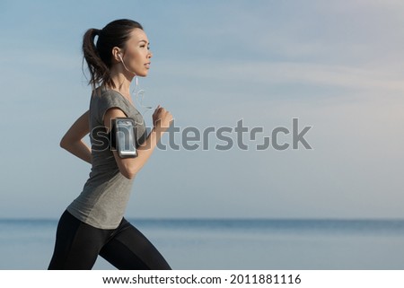Outdoor shot of young sports asian woman running along the sea shore in morning. Female athlete jogging in sporty fitness clothes for shaping her body, weightloss