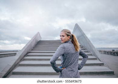 Outdoor Shot Of Young Woman Standing By A Stairs And Looking Over Shoulder. Fitness Female Model Ready For A Run.