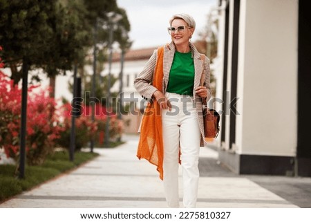 Outdoor spring fashion portrait of beautiful mature woman wearing stylish clothes posing in city street and smiling. Happy model enjoying life