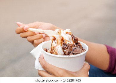 Outdoor Street Closeup View Of Hand Hold A Cup, Plastic Spoon, And Eat Melting Frozen Yogurt Ice Cream With Chocolate Topping In White Paper Cup.