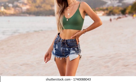 Outdoor summer image of sexy sportive blond woman posing on the beach. Wearing jeans shorts and green top. Fashion details.