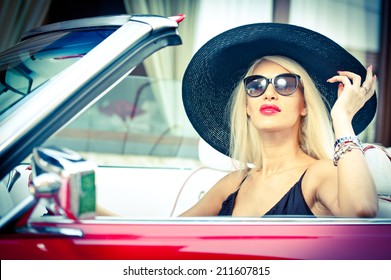 Outdoor Summer Portrait Of Stylish Blonde Vintage Woman Driving A Convertible Red Retro Car. Fashionable Attractive Fair Hair Female With Black Hat In Red Vehicle. Sunny Bright Colors, Outdoors Shot.