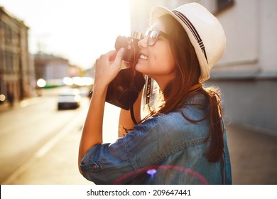 Outdoor summer smiling lifestyle portrait of pretty young woman having fun in the city in Europe in evening with camera travel photo of photographer Making pictures in hipster style glasses and hat 