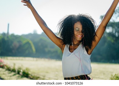 Outdoor sunny portrait of the adorable active african girl with green eye shadows and pretty smile dancing and enjoying music in earphones.