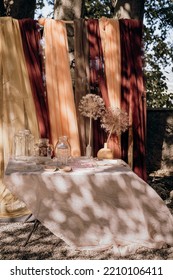 Outdoor Table Dressed With Lightweight Fabrics In Apricot And Burgundy Tones. Dappled Light On A Warm Breezy Afternoon. Empty Glass Jars And Plates Waiting To Be Filled By Caterer For Outdoor Event.