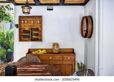 Outdoor terrace wine corner with wine bottle and glasses wooden holder on the white wall,  fruits and vase with winecorks on cupboard, part of round table. Selective focus
