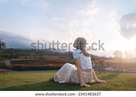 Outdoor travel in nature concept. Rear view of woman relax sitting in botanical garden flower blossom park field with sunlight. Background with mountain and sky.