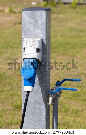 An outdoor utility post featuring a blue industrial electrical plug connected to a weathered socket and two water faucets, ideal for camping, RV hookup, or utility service in a grassy setting.