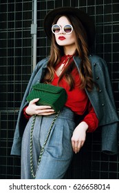 Outdoor Waist Up Portrait Of Beautiful Woman Posing On Street. Model Wearing Stylish Red, Grey Clothes, Wide-brimmed Hat, Round Sunglasses, Holding Green Quilted Suede Bag. Female Fashion Concept