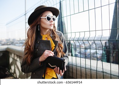 Outdoor waist up portrait of young beautiful woman with long hair, looking aside. Model wearing stylish hat,  white round sunglasses, clothes, holding small bag. City lifestyle. Female fashion concept