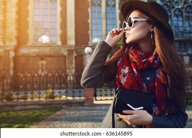 Outdoor Waist Up Portrait Of Young Beautiful Woman Posing On Street. Model Looking Aside, Wearing Stylish Hat, Sunglasses, Scarf, Holding Small Bag. Sunset. Female Fashion. Copy, Empty Space For Text