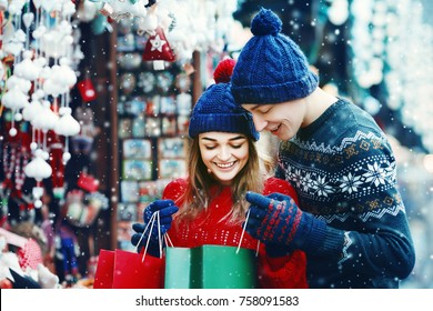 Outdoor Waist Up Portrait Of Young Beautiful Happy Smiling Couple Holding And Looking At Bags. Shopping At The Street Christmas Fair. Models Wearing Stylish Winter Clothes. Snowfall. Copy, Empty Space