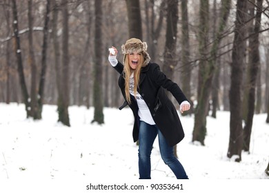 Outdoor Winter Portrait Of A Beautiful Happy Blond Young Woman In A Snowball Fight In The Woods. She Is Smiling, Wearing A Winter Hat, And Holding A Snowball. Very Shallow Depth Of Field.