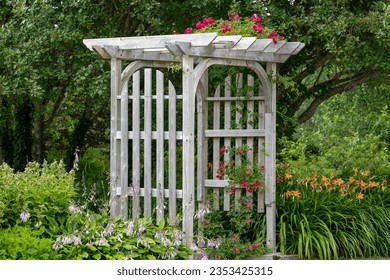 An outdoor wooden curved shaped archway or arbor surrounded by a lush green garden.  The park has birch trees, climbing red roses, orange lily flowers, and vibrant green shrubs in a botanical park.  - Shutterstock ID 2353425315