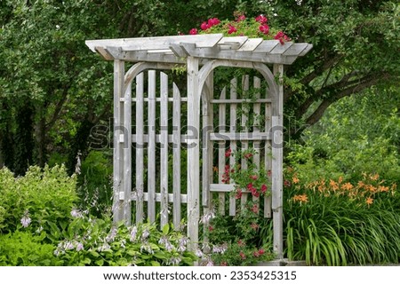 An outdoor wooden curved shaped archway or arbor surrounded by a lush green garden.  The park has birch trees, climbing red roses, orange lily flowers, and vibrant green shrubs in a botanical park. 