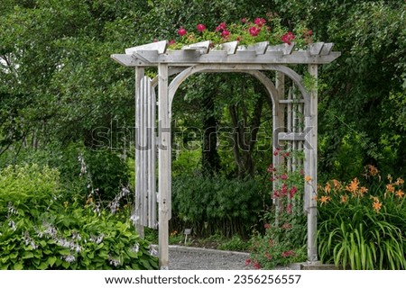 An outdoor wooden curved shaped archway or arbor surrounded by a lush green garden.  The park has birch trees, climbing red roses, orange lily flowers, and vibrant green shrubs in a botanical park. 