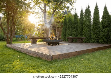 An outdoor wooden deck features a circular fire pit with burning logs. Surrounded by lush greenery and tall evergreens, rustic benches enhance the serene setting. - Powered by Shutterstock