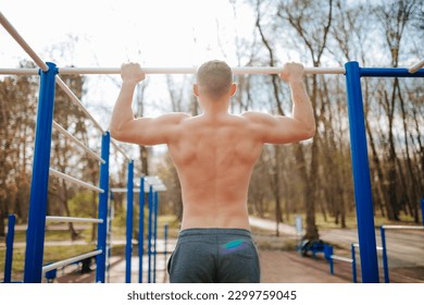 Outdoor Workout Guy Performs Intense Pull-Ups on a Bar. A fit male trains his upper body by doing naked pull-ups on a bar outside, with no shirt to hold him back.