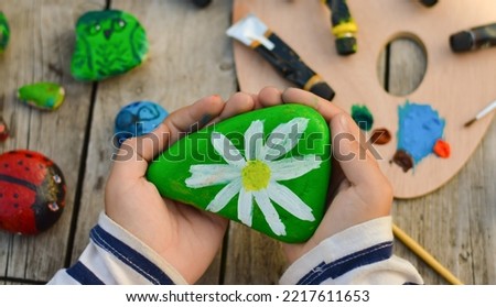 Outdoor workshop in summer. A selective focus of a schoolboy boy holding a painted flat stone in his hands. Do it yourself.