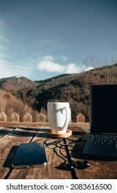 Outdoor Workspace With Laptop, Cup Of Coffee, Smartphone And Earphones On A Wooden Table In The Mountain