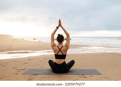 Outdoor yoga. Woman meditating with hands clasped above head, sitting in lotus position on fitness mat on beach, practicing meditation near ocean, back view