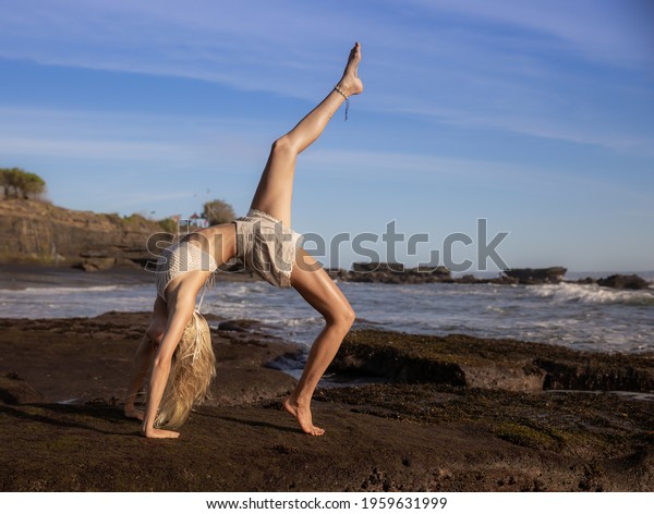 Outdoor yoga. Young woman practicing Eka Pada\
Chakrasana, One Legged Wheel Pose. Upward facing bow pose is a deep\
backbend. Flexible, fit body. Yoga retreat. Copy space. Beach in\
Bali, Indonesia