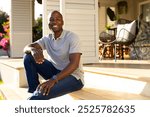 Outdoors, African American young man sitting on porch steps, smiling at camera. Sunlit patio with outdoor furniture and plants in background, creating warm and inviting atmosphere, unaltered