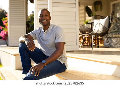 Outdoors, African American young man sitting on porch steps, smiling at camera. Sunlit patio with outdoor furniture and plants in background, creating warm and inviting atmosphere, unaltered - Powered by Shutterstock