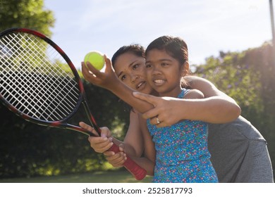 Outdoors biracial mother and daughter teaching tennis, focused on holding racket. They are outdoors on sunny day with green background, unaltered - Powered by Shutterstock