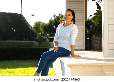 Outdoors, biracial woman in her late thirties sitting on porch steps, smiling warmly. Enjoying sunny day in well-maintained garden with lush greenery and white background house, unaltered - Powered by Shutterstock