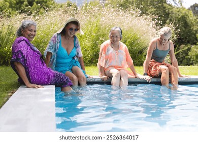 Outdoors, diverse senior female friends sitting by pool, dipping feet in water, copy space. Wearing colorful summer clothes, sharing joyful moments together, enjoying sunny day, unaltered - Powered by Shutterstock