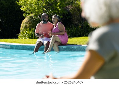 Outdoors, diverse senior friends, laughing by pool, holding drinks. An African American man in orange shirt and biracial woman in pink dress, enjoying sunny day, unaltered - Powered by Shutterstock