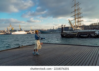 Outdoors Fashion Portrait Of A Beautiful Middle Aged Woman Walking On The Beach. Marine Background. Dressed In A Stylish Warm Blue Sweater, Yellow Skirt And Beret.