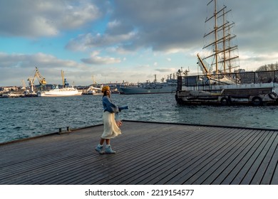 Outdoors Fashion Portrait Of A Beautiful Middle Aged Woman Walking On The Beach. Marine Background. Dressed In A Stylish Warm Blue Sweater, Yellow Skirt And Beret.