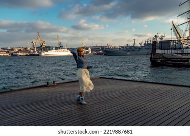 Outdoors Fashion Portrait Of A Beautiful Middle Aged Woman Walking On The Beach. Marine Background. Dressed In A Stylish Warm Blue Sweater, Yellow Skirt And Beret.