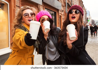 Outdoors fashion portrait of company cheerful pretty girls friends drinking coffee. Walking in the city. Talking and going shopping. Wearing stylish outerwear, sunglasses and hats. Bright make up