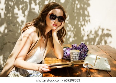 Outdoors fashion portrait of stunning woman sitting in cafe. Drinking coffee and reading old book. Wearing stylish long beige waistcoat, striped skirt and vintage sunglasses. Close up. Get inspired