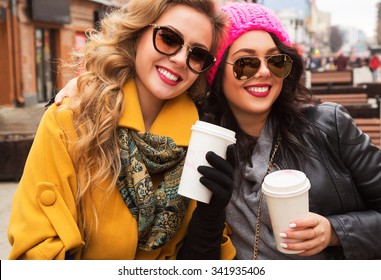 Outdoors fashion portrait of two cheerful pretty girls friends walking in the city. Talking, drinking coffee and going shopping. Wearing stylish outerwear, hats and sunglasses. Bright make up 