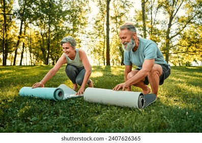 Outdoors fitness of couple. Sporty family of two old people with bare feet laying yoga mats on grass to exercise on fresh air. Retired man and woman preparing for joint yoga practice at sunny park. - Powered by Shutterstock