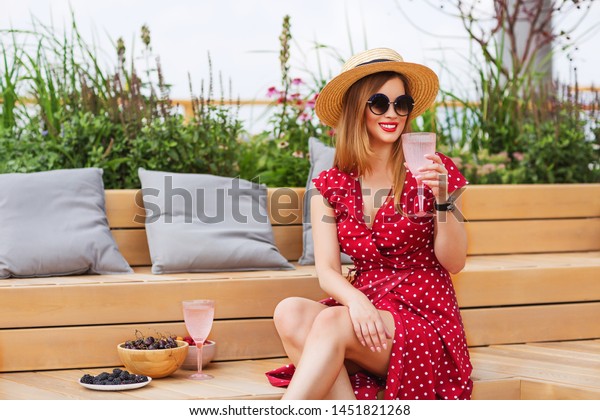 Outdoors lifestyle fashion portrait of\
beautiful young women sitting on the patio. Drinking champagne,\
eating berries, smiling and enjoying life. Wearing stylish dress\
polka dots, canotier,\
sunglasses