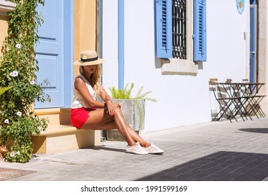 Outdoors lifestyle fashion portrait of pretty young woman sitting on stoop. Wearing stylish straw hat, sunglasses and red shorts. Travelling, walking on the summer city. Summer holiday. Relaxation