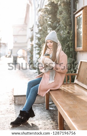 Outdoors lifestyle fashion portrait of pretty blonde girl walking on the holiday city. Smiling, drinking coffee. Wearing stylish pink coat and wide scarf. Christmas mood