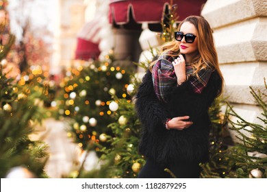 Outdoors Lifestyle Fashion Portrait Of Stunning Blonde Girl. Smiling, Walking On The Christmas City. Going Shopping. Trendsetter. Wearing Stylish Black Fur Coat, Scarf And Sunglasses. Festive Mood